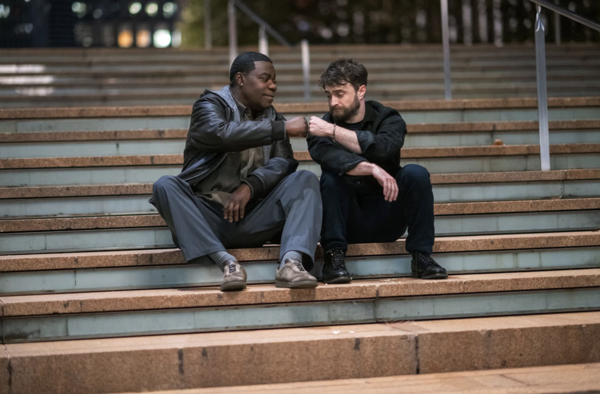 A Black man and white man sit on a long set of outdoor steps. They are bumping fists.