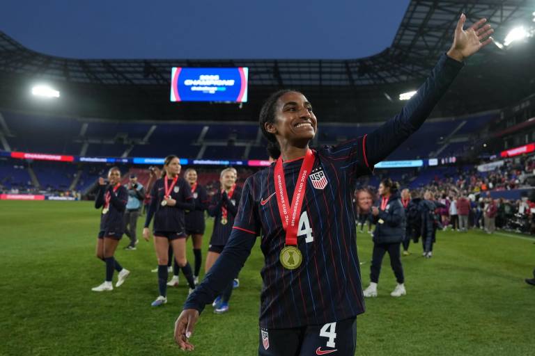 A woman in a soccer jersey and a gold medal waves at fans as she walks off of the soccer pitch.