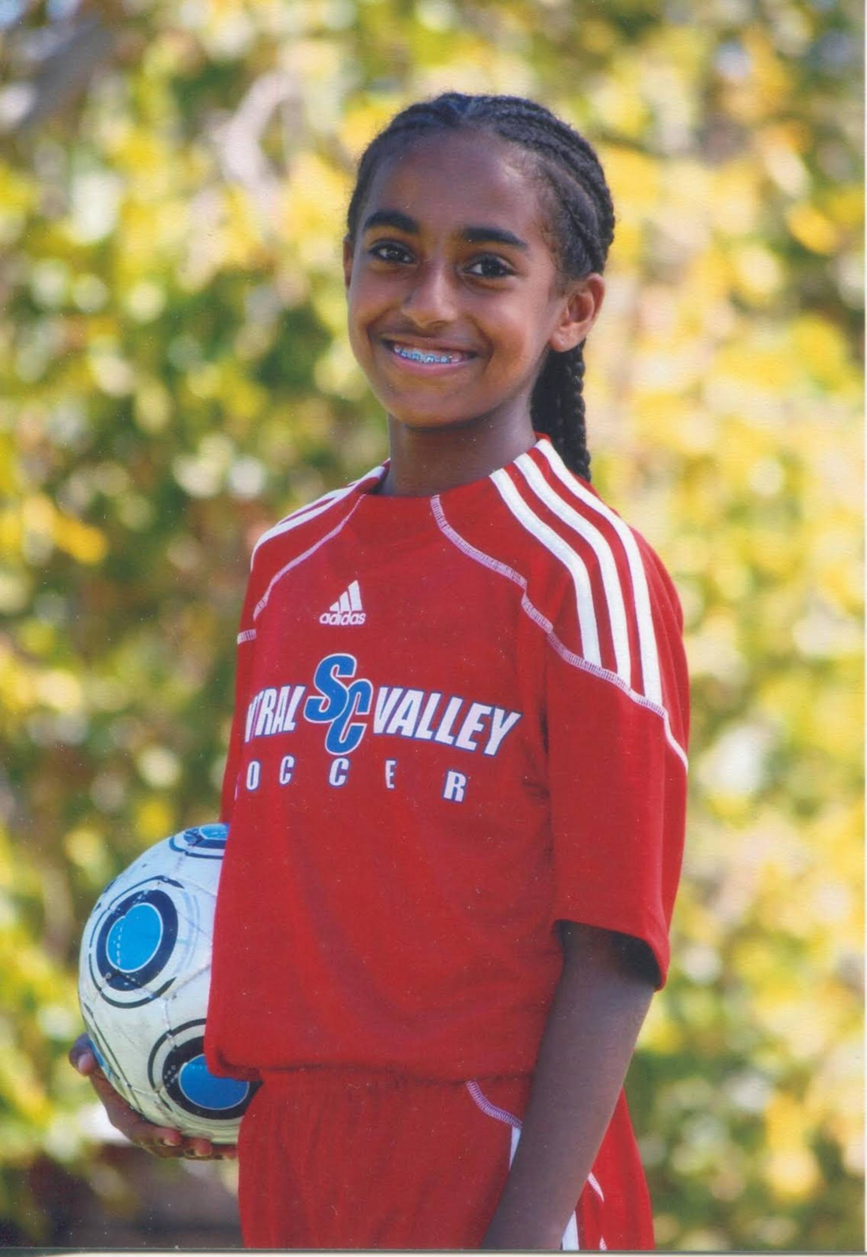 A young girl in a red soccer jersey, posing for a photo while holding a soccer ball. 