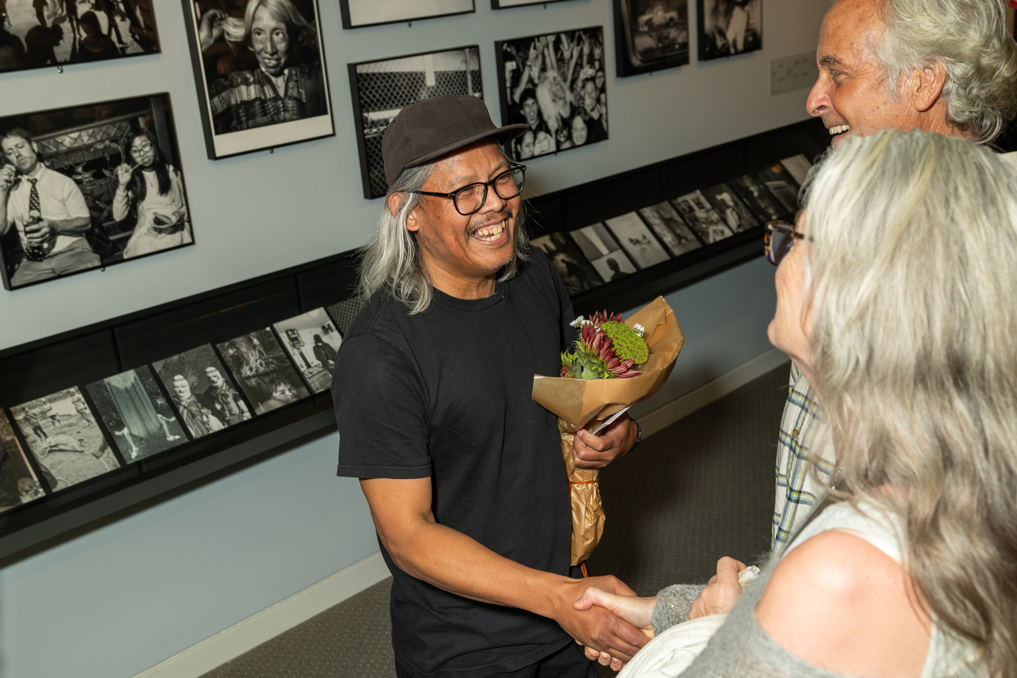 An Asian man with long grey hair smiles, wearing a Black t-shirt, black baseball cap and black rimmed glasses. He is holding a bunch of flowers and standing in front of a wall full of framed black and white photography.