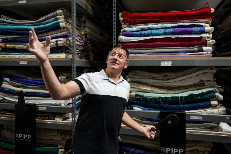 while man gestures upward, folded stacks of quilts behind him