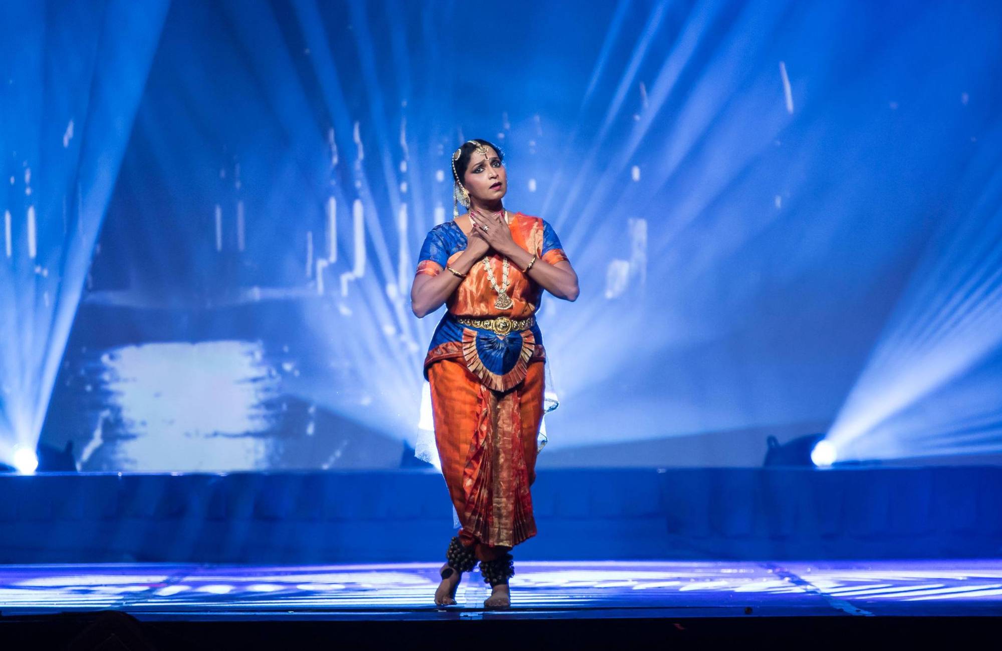 An Indian woman in colorful wardrobe holds her hands against her chest and looks outward against a blue lit background.