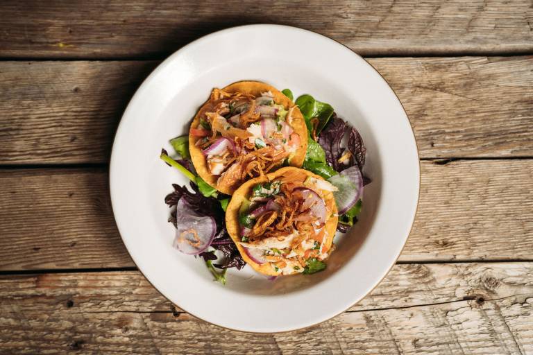 Overhead view of a plate of two tostadas topped with fish and fried shallots, served on a wooden picnic table.