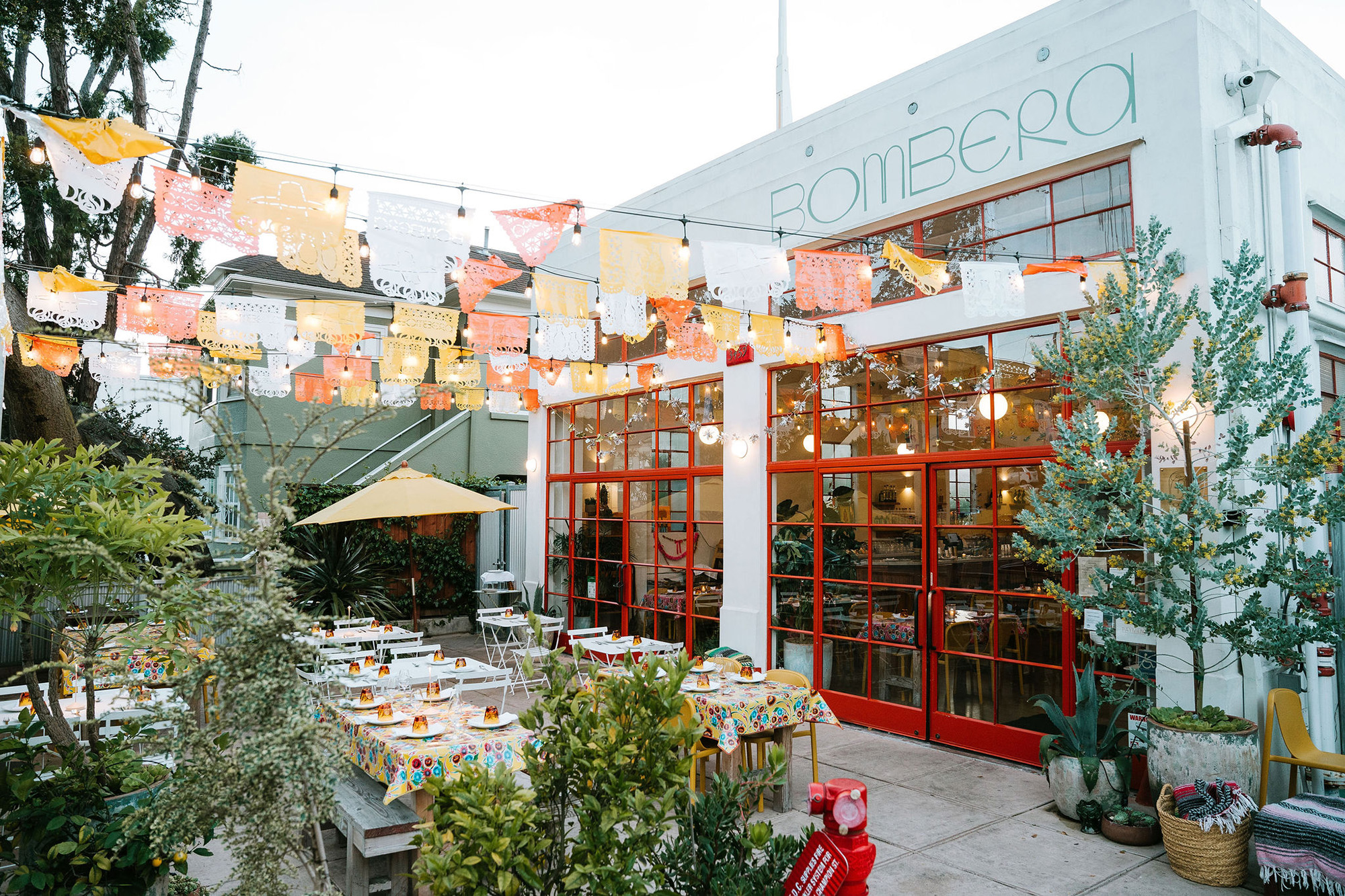 View of a restaurant courtyard decorated with festive banners. The name of the restaurant, "Bombera," is visible on its facade.