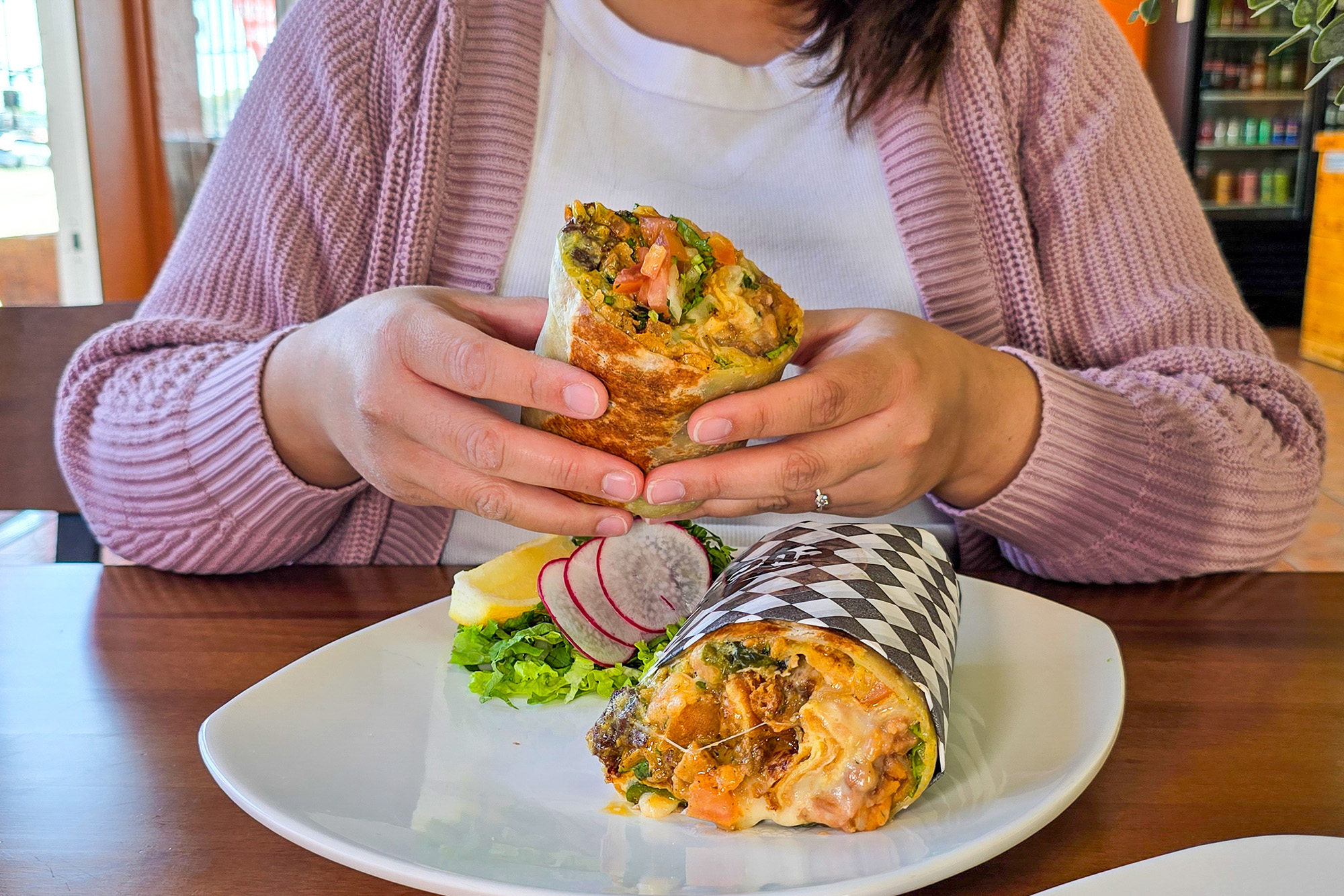 A woman in a pink cardigan holds a burrito in her hand, ready to take a bite.