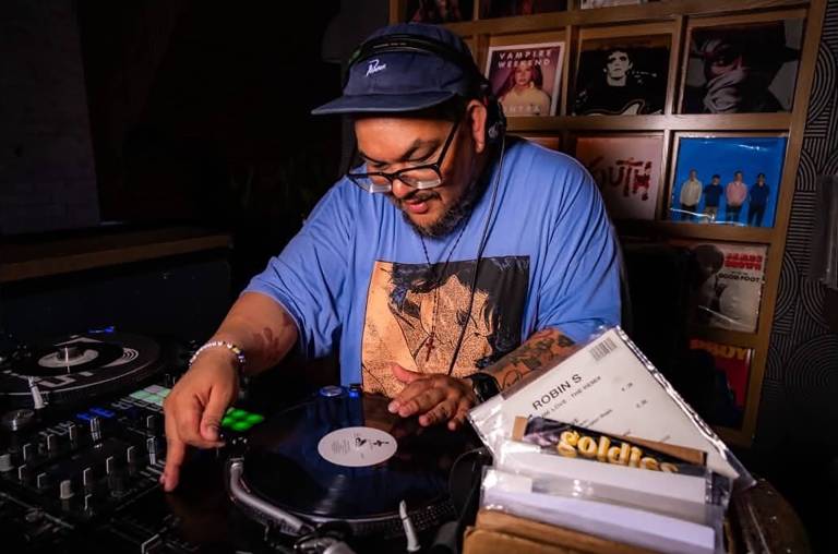 A Latino DJ in a blue shirt and cap works the turntables, looking downward.