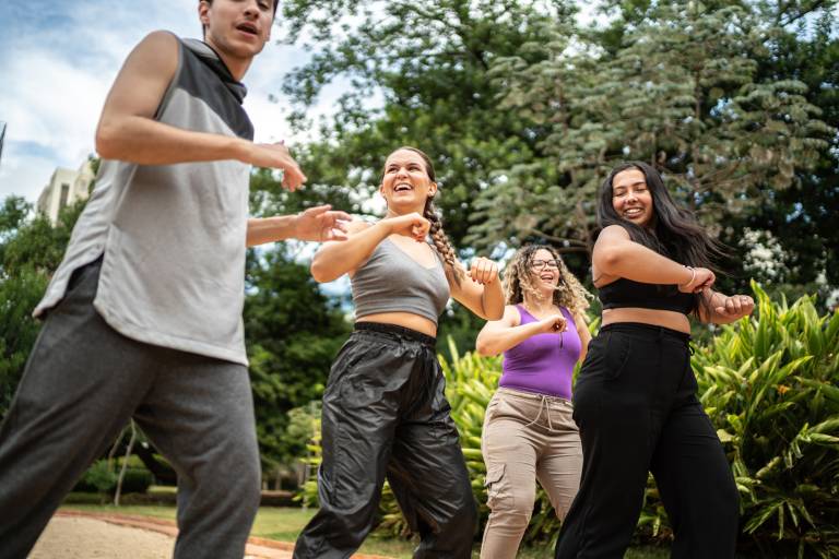 A group of people of various ethnicities dances outdoors against the trees and bushes of a park