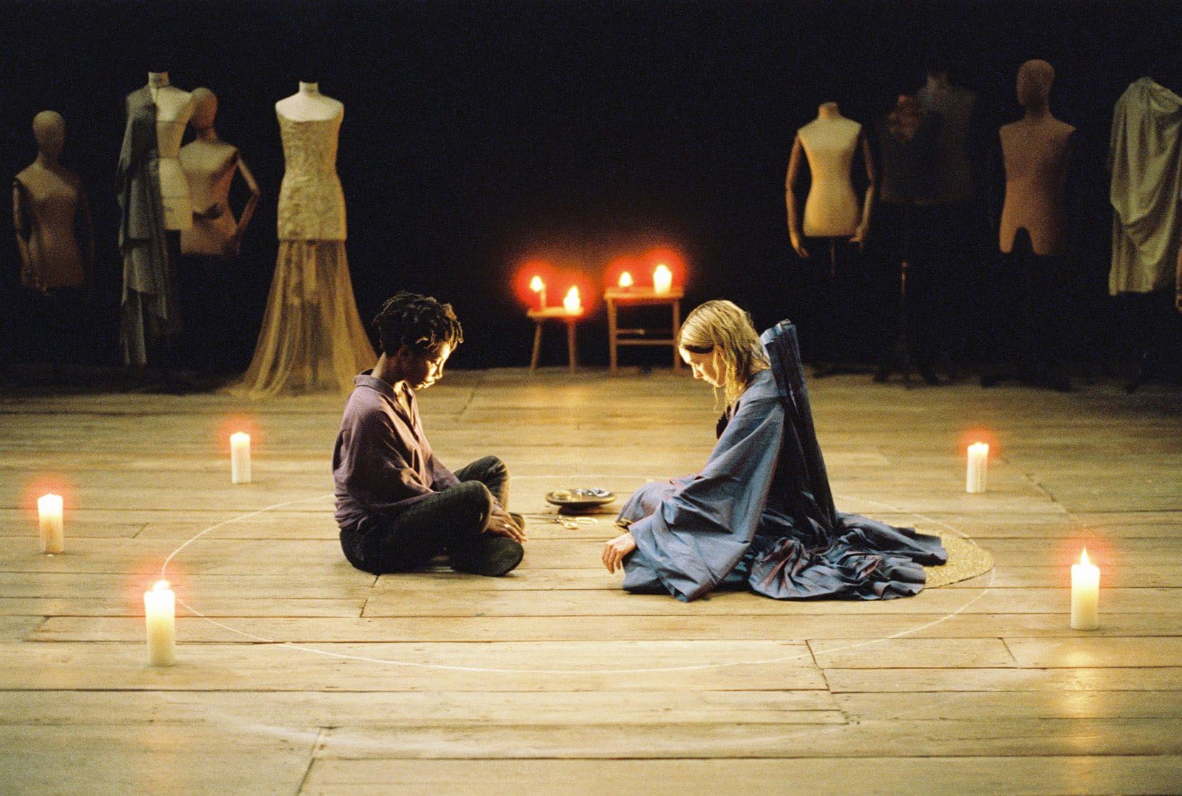 Two women sit cross-legged, facing each other, surrounded by candles and mannequins.