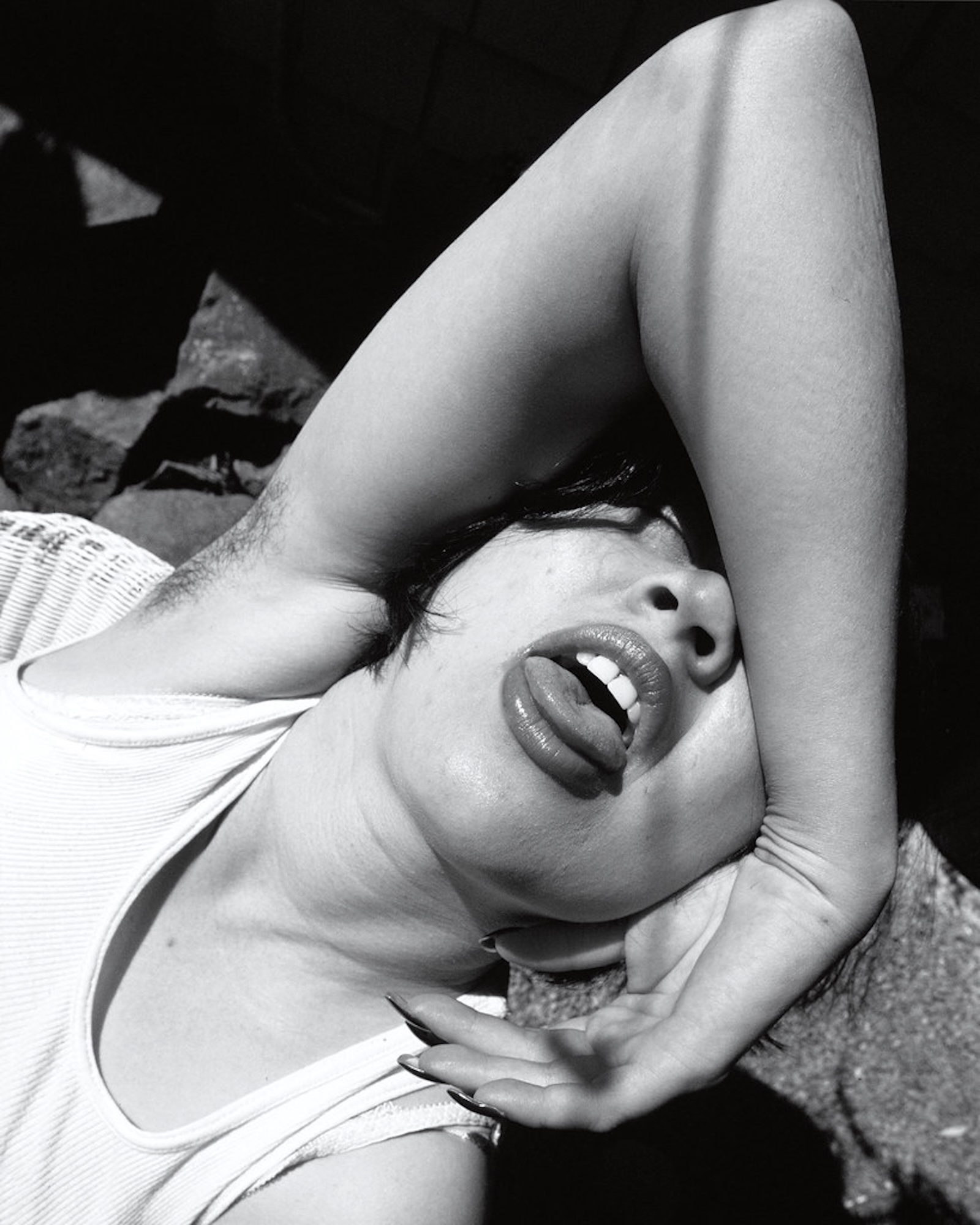 A woman lies with her upper face obscured by her own arm which is raised to reveal a hairy armpit. She sticks her tongue out.