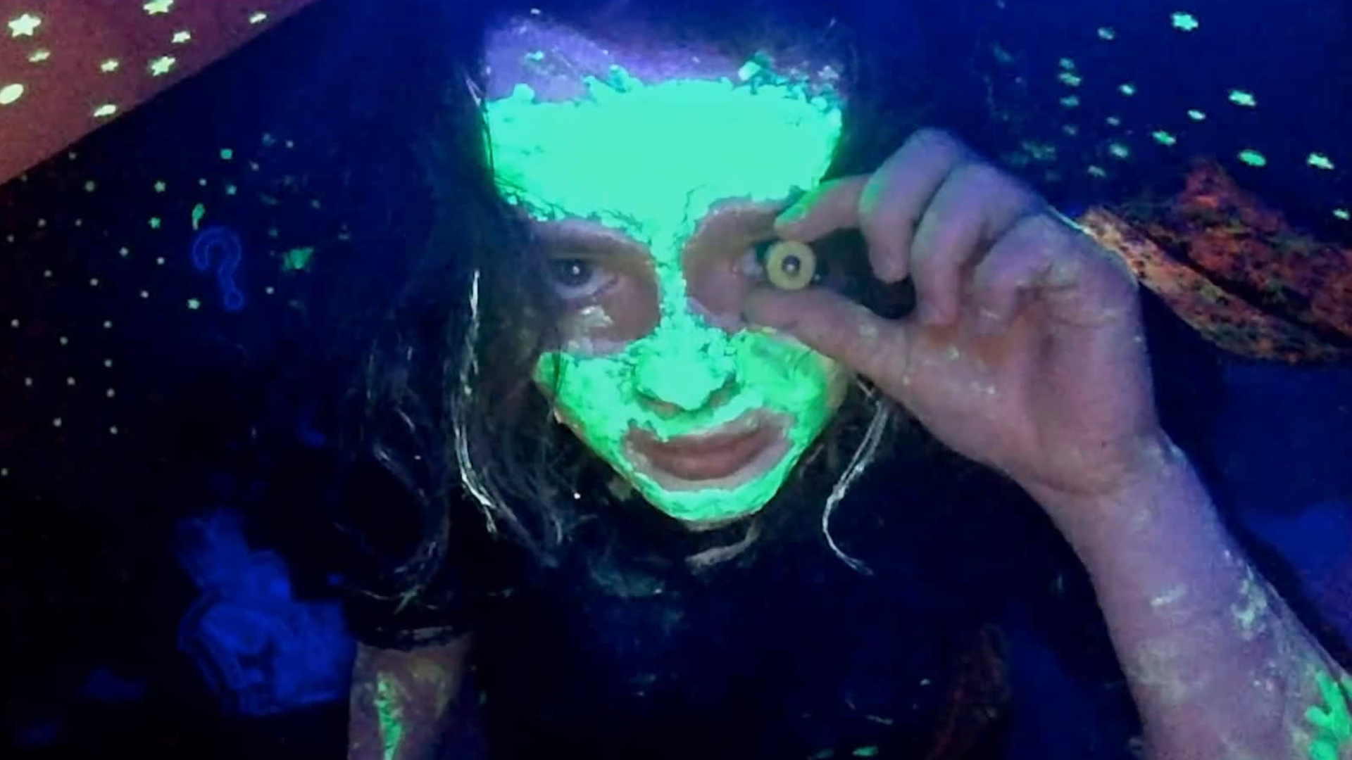 person with glowing face paint holds a fake eye to their own eye