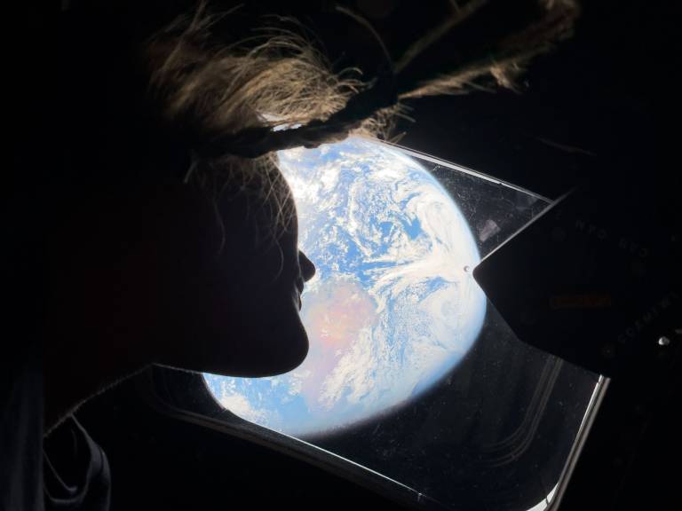 A woman's hair and face is seen in silhouette next to a spacecraft's window, looking at a view of the Earth from a great height.