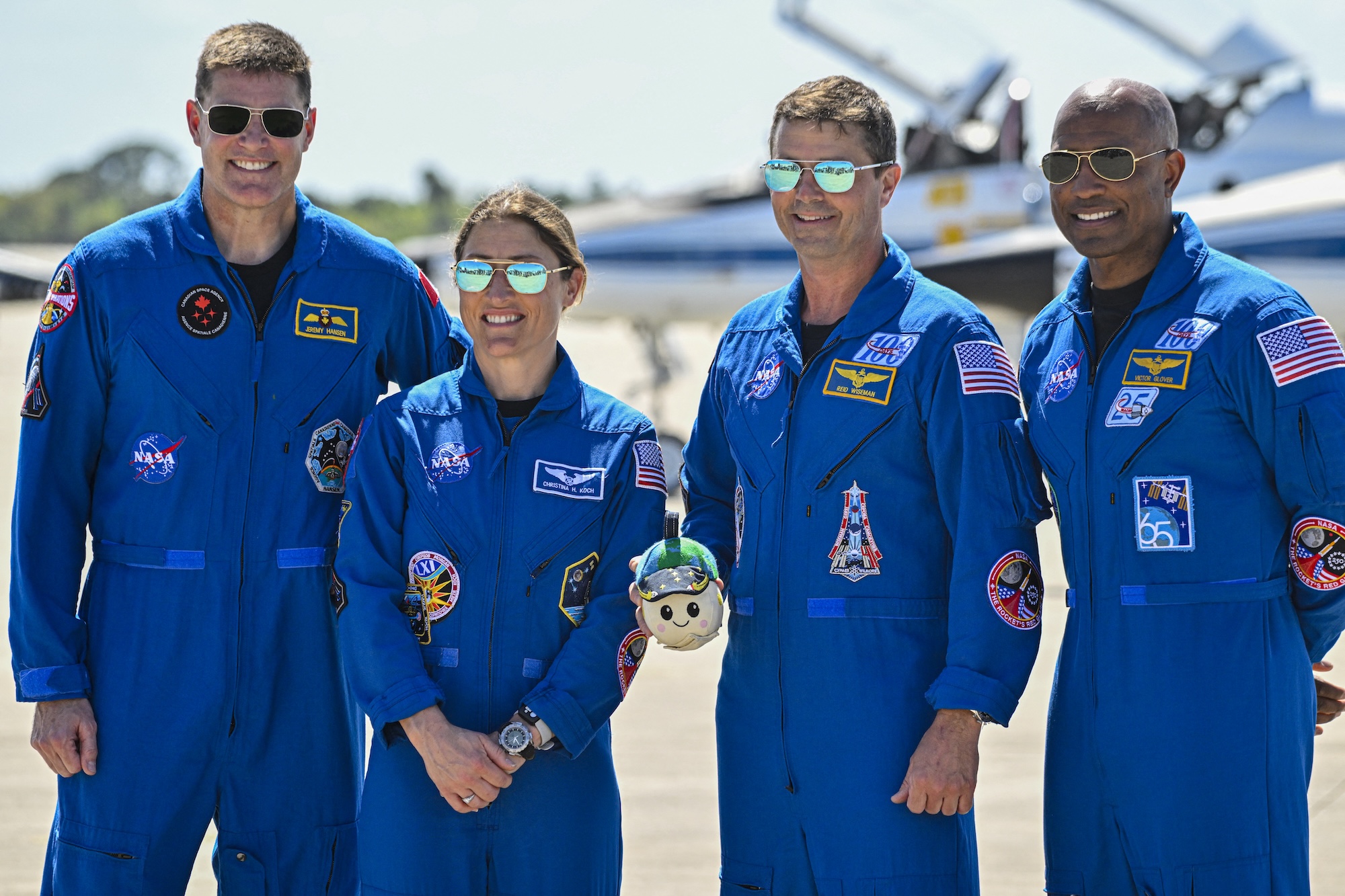 Two white men, a white woman and a Black man stand in a line smiling, all wearing matching blue jumpsuits and aviator sunglasses on tarmac on a sunny day.