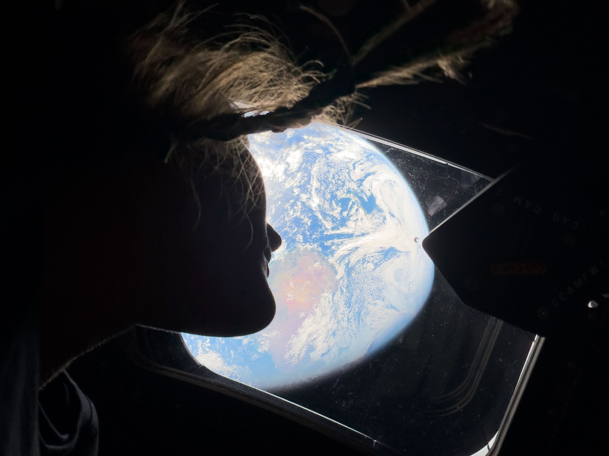A woman's hair and face is seen in silhouette next to a spacecraft's window, looking at a view of the Earth from a great height.