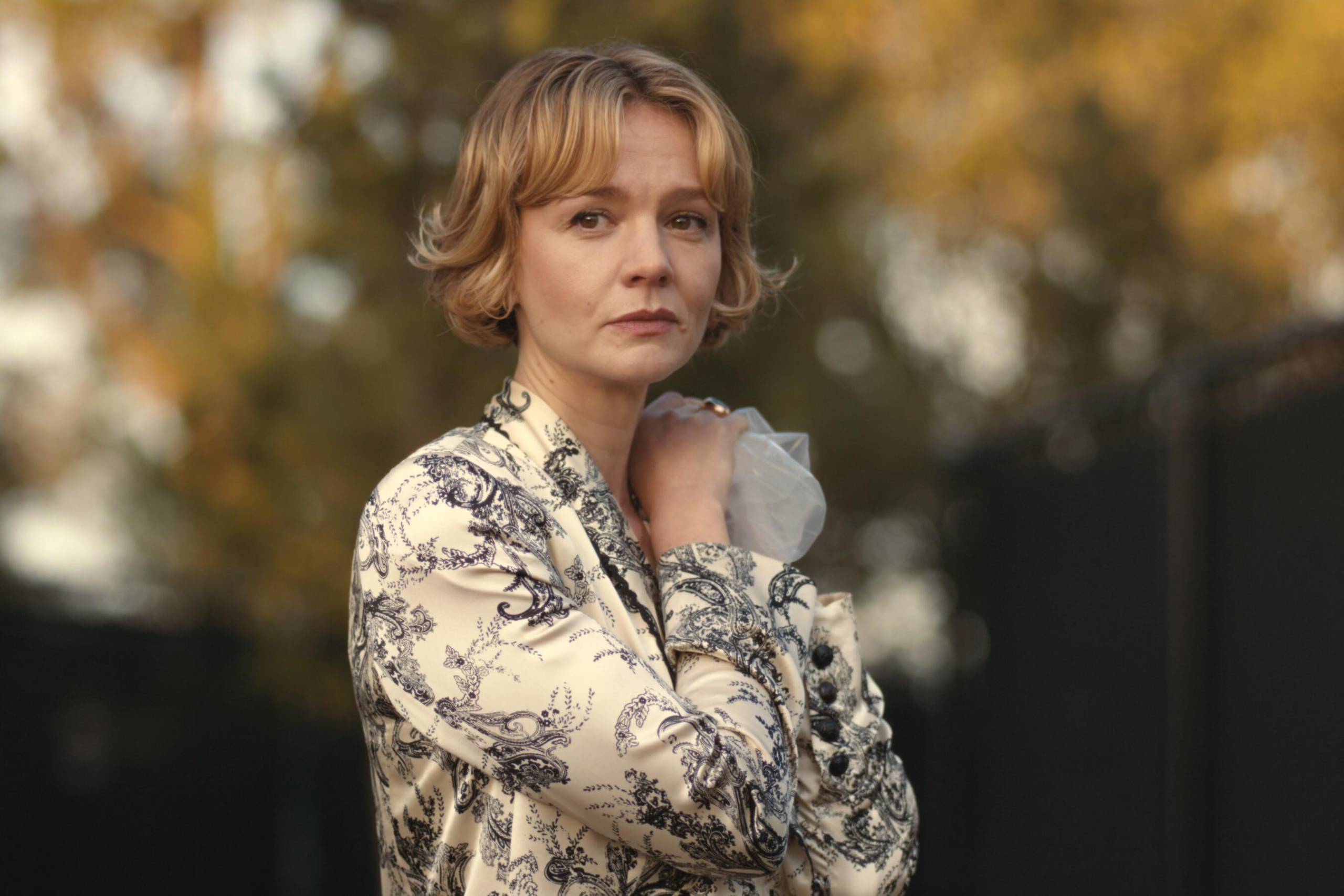A petite middle aged white woman with short blond hair stands outdoors, looking wistfully into the distance.