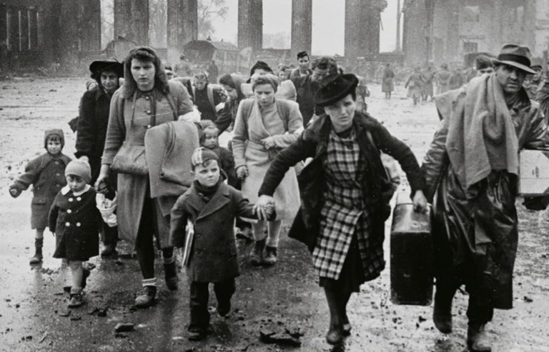 A black and white photo of men. women and children in Berlin in the 1940s, carrying suitcases and fleeing the city on foot.