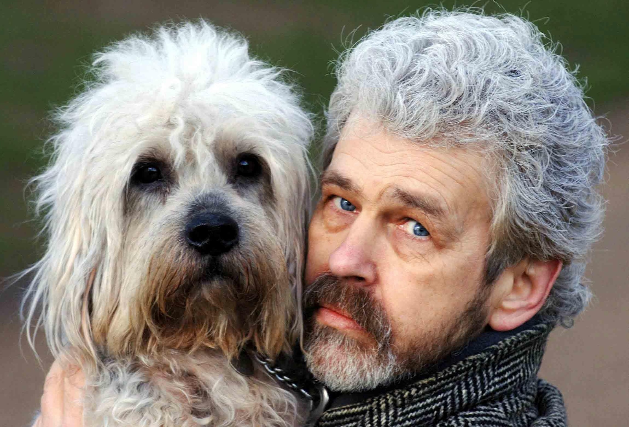 A shaggy white dog with dark eyes poses face to face with his owner, who has similar white hair and dark eyes. 