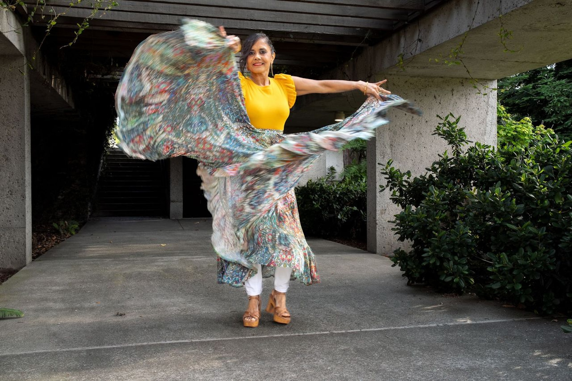A woman in a bright top and colorful skirt dances as she takes a photo.