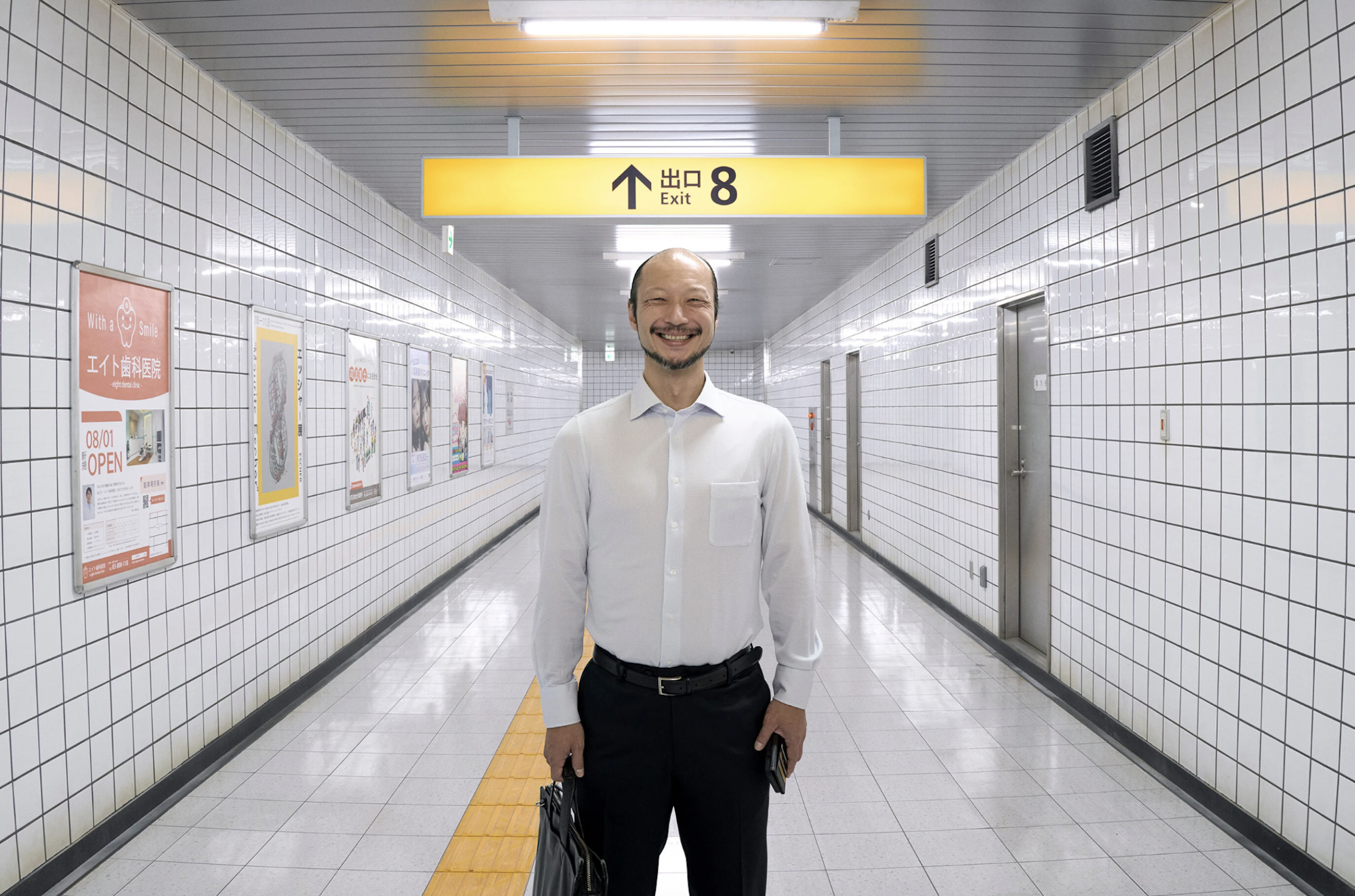 A Japanese man, wearing a white shirt and black pants stands in the center of a brightly lit underground corridor. He is smiling unnaturally.