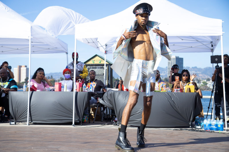 A slender Black man poses before a panel of judges wearing a leather hat and boots, and matching holographic shorts and open jacket. It's a sunny day.