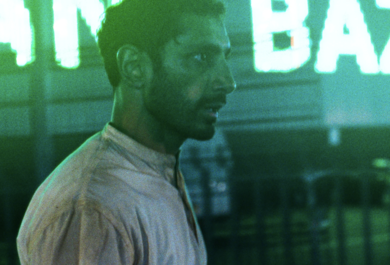 A man of Pakistani descent, in his thirties, walks through neon-lit streets, looking worse for wear.