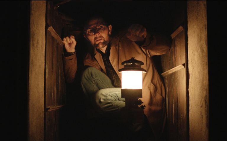 A white middle aged man with glasses crouches inside a wall crawl space clutching a lit lantern. He is dressed in casual jeans, plaid shirt and tan jacket.