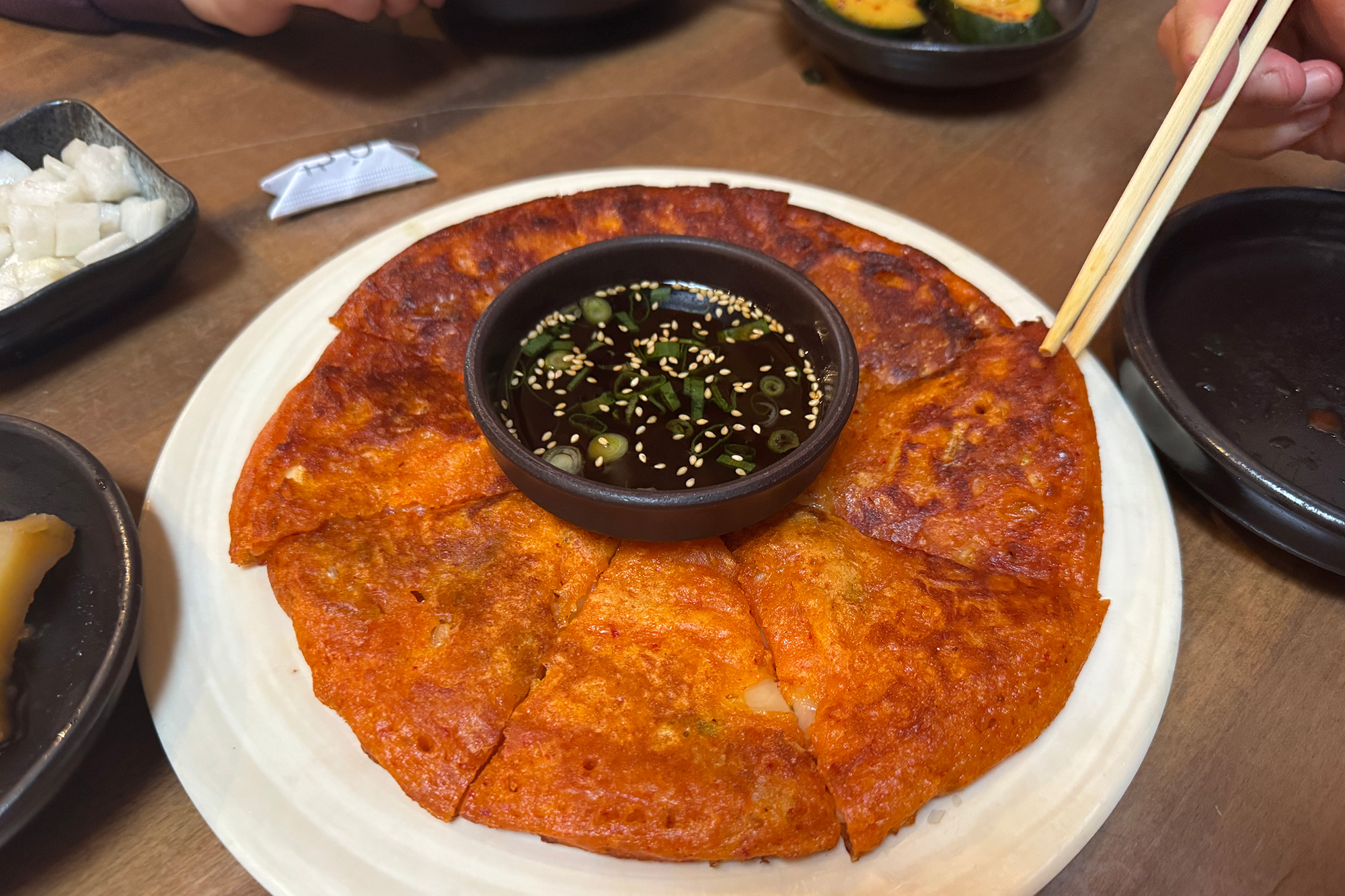 A very crispy-looking, orange-tinted kimchi pancake, cut in wedges on a plate.