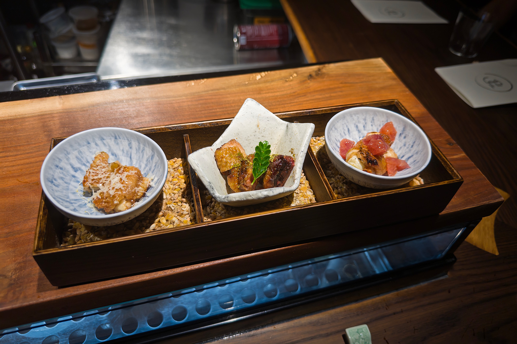 Three small plates of yakitori, presented in an elegant wooden box.