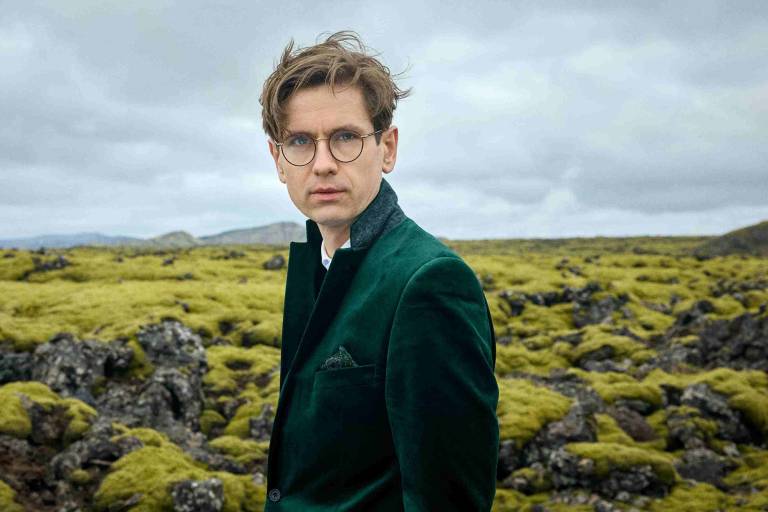 A 42-year-old man in a deep green jacket stands against a cloudy sky and lush green fields and rocks.
