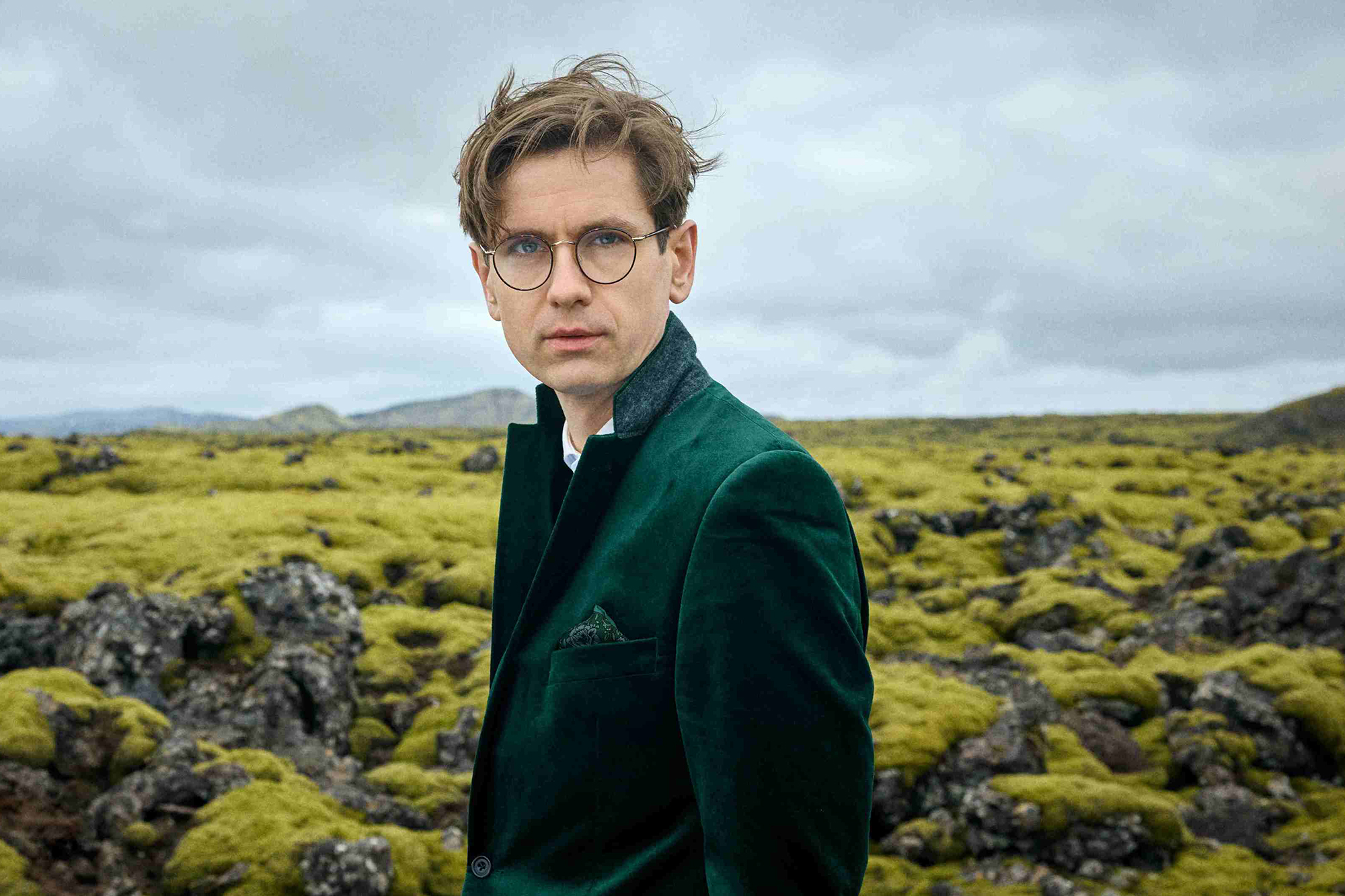 A 42-year-old man in a deep green jacket stands against a cloudy sky and lush green fields and rocks.