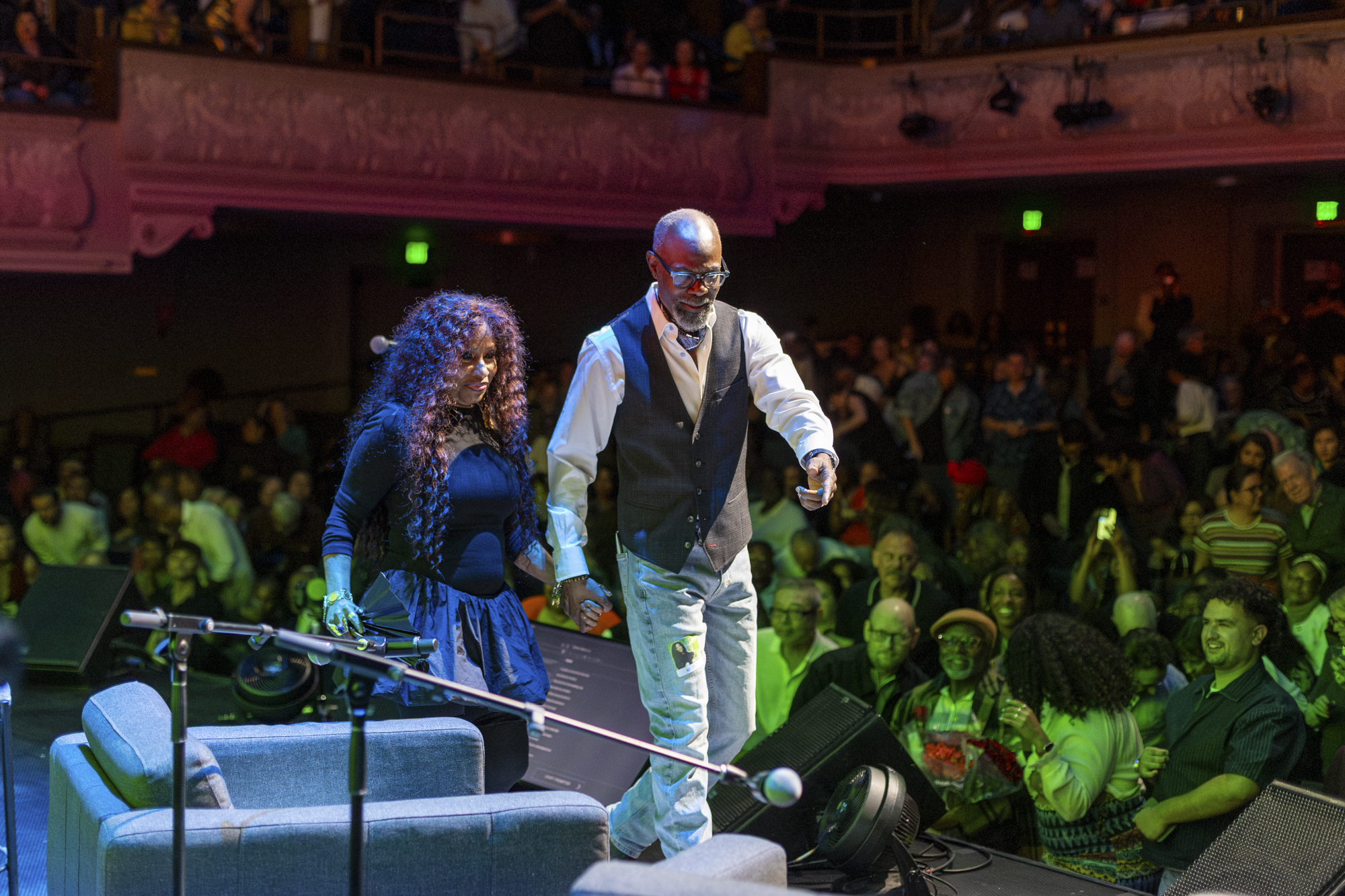 Black woman and Black man stride across stage in front of large audience