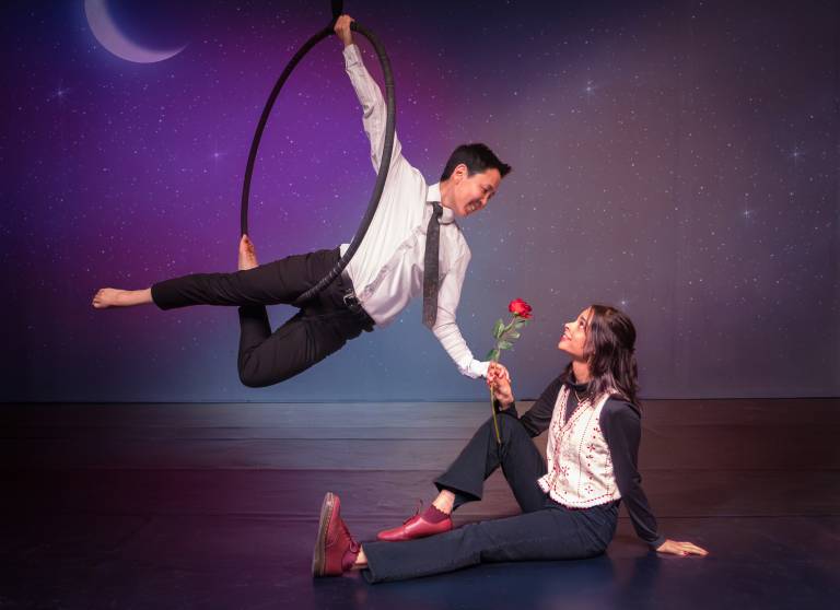 A young man srtiking a cupid-like pose in a suspended hoop hands a rose to a young attractive woman sitting on the floor of a stage