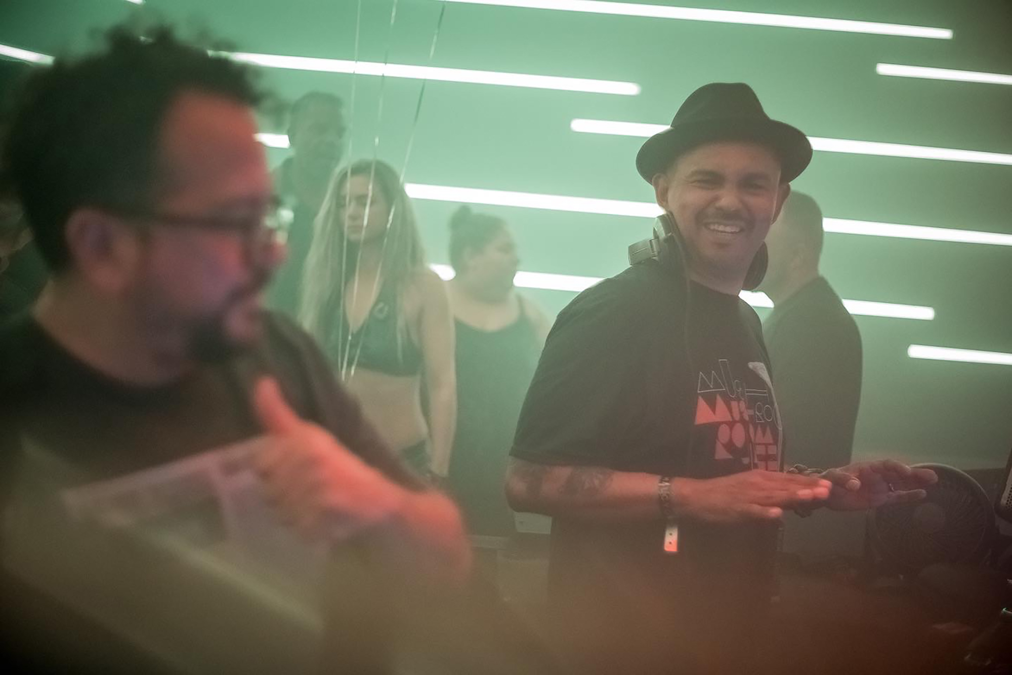 Two men in their 30s-40s stand at a DJ table, against a fuzzy background of horizontal green lighting