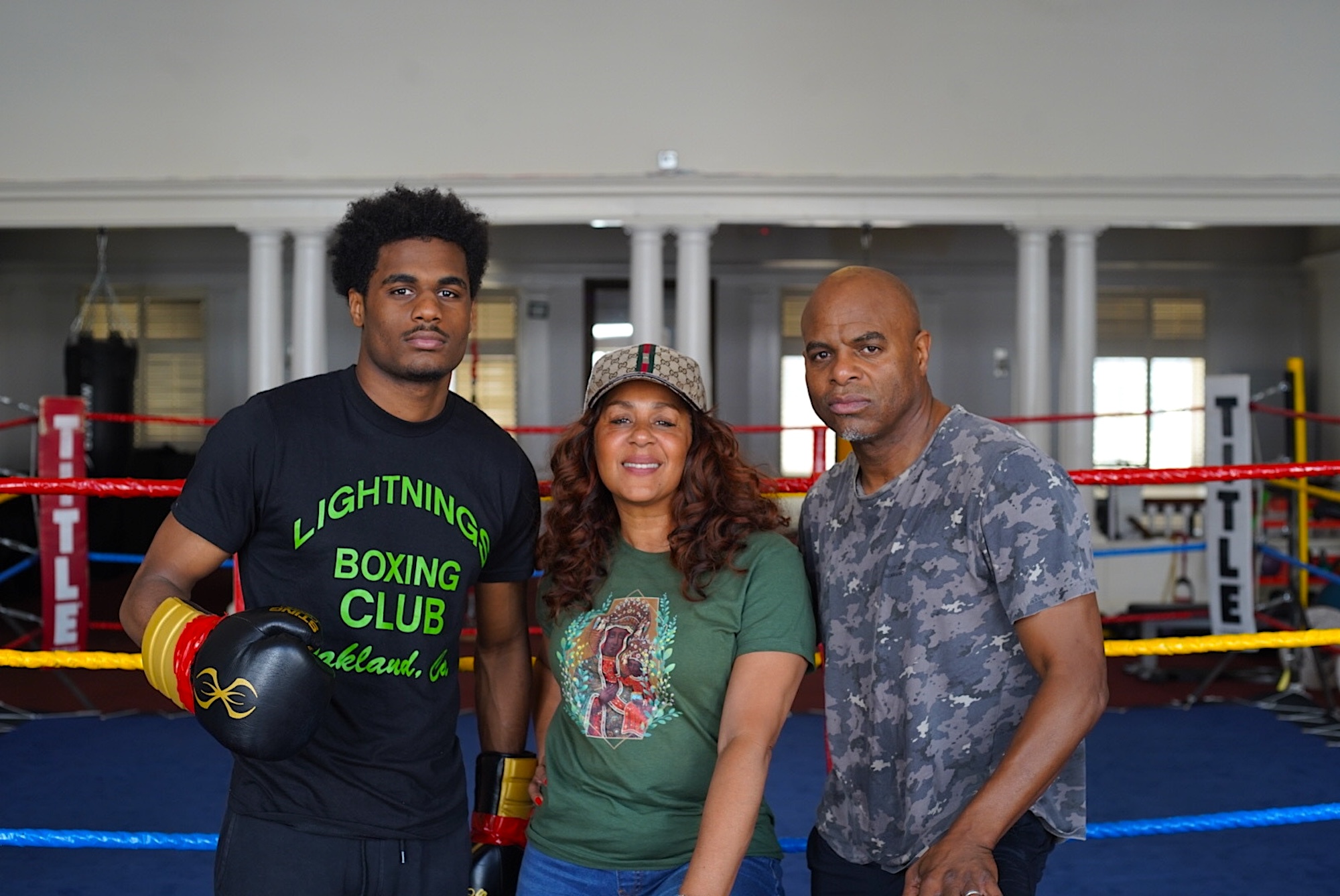 A young African American man wears boxing gloves as he poses for a photo next to his mother and father. 