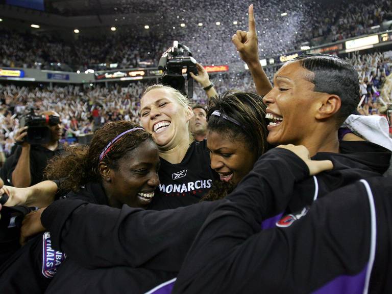 A group of women in tracksuits joins in a group hug as confetti falls from the ceiling of an arena