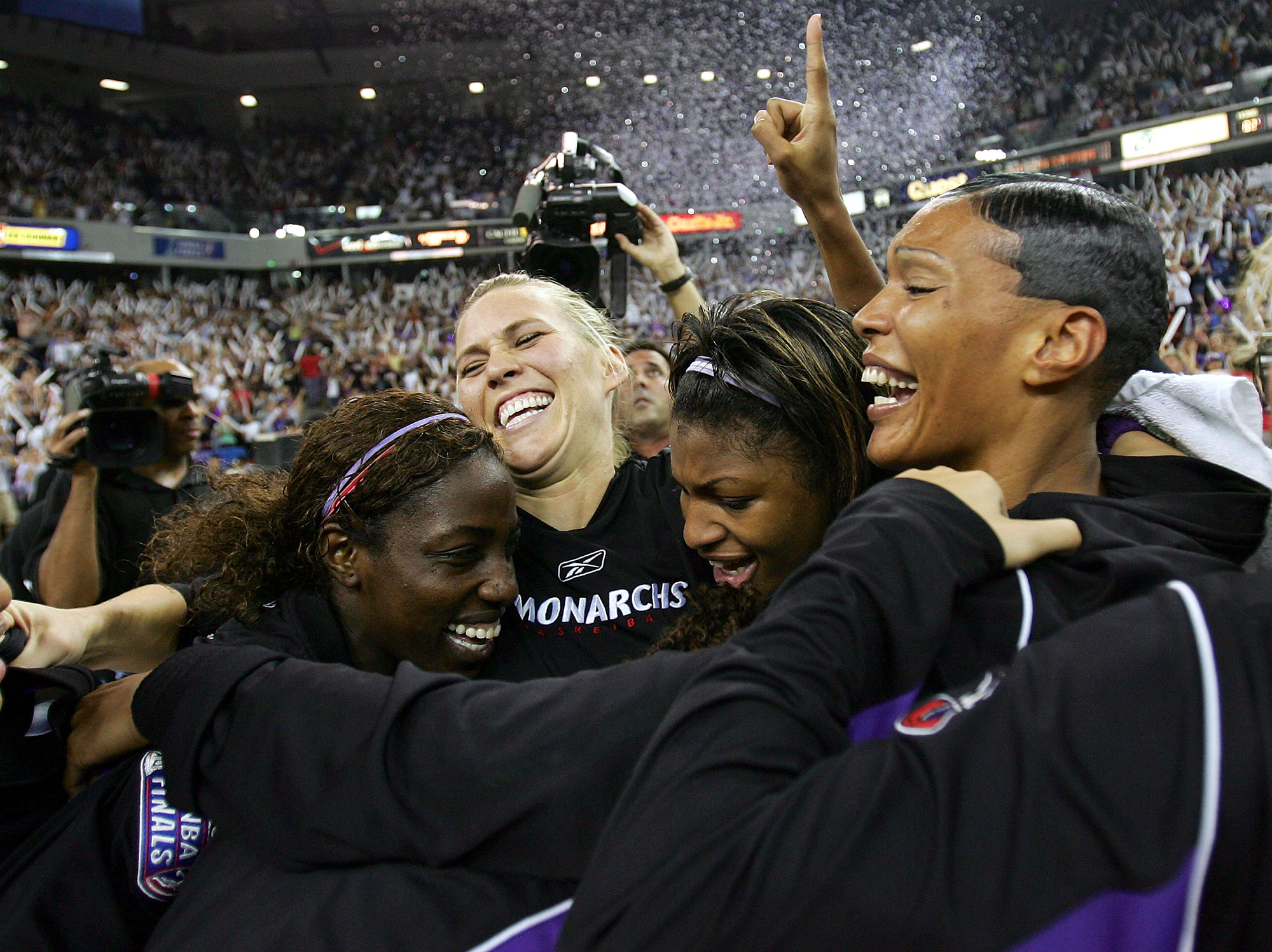 A group of women in tracksuits joins in a group hug as confetti falls from the ceiling of an arena