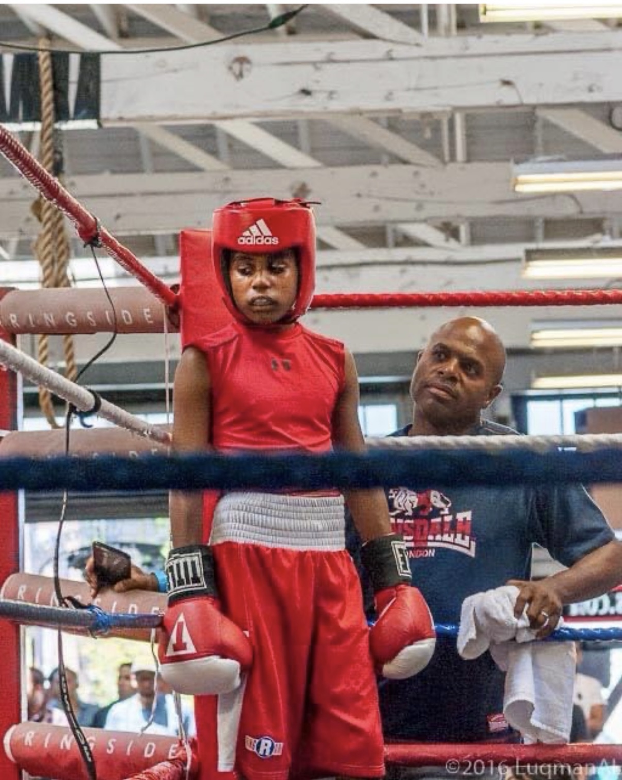 A boy stands in a boxing ring, as his father stands outside of the ring looking on in admiration. 