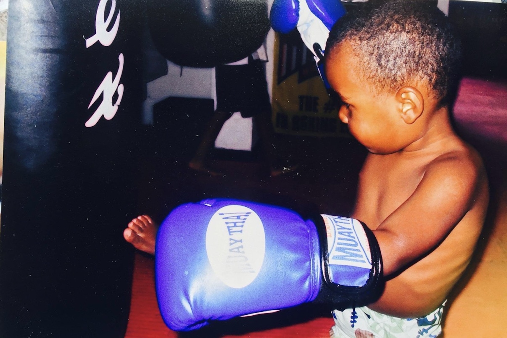 An African American toddler wearing boxing gloves and kicking a punching bag. 