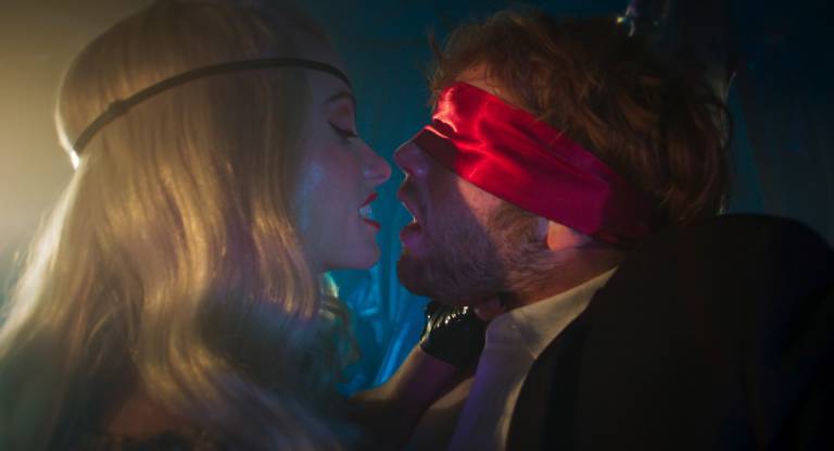 blonde woman and blindfolded man face each other in near kiss