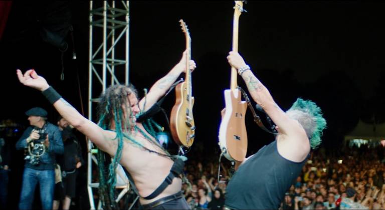On stage, before a huge audience, a shirtless white man with green dreadlocks faces a white man with a green mohawk. Both gesture at each other wildly, holding guitars aloft.