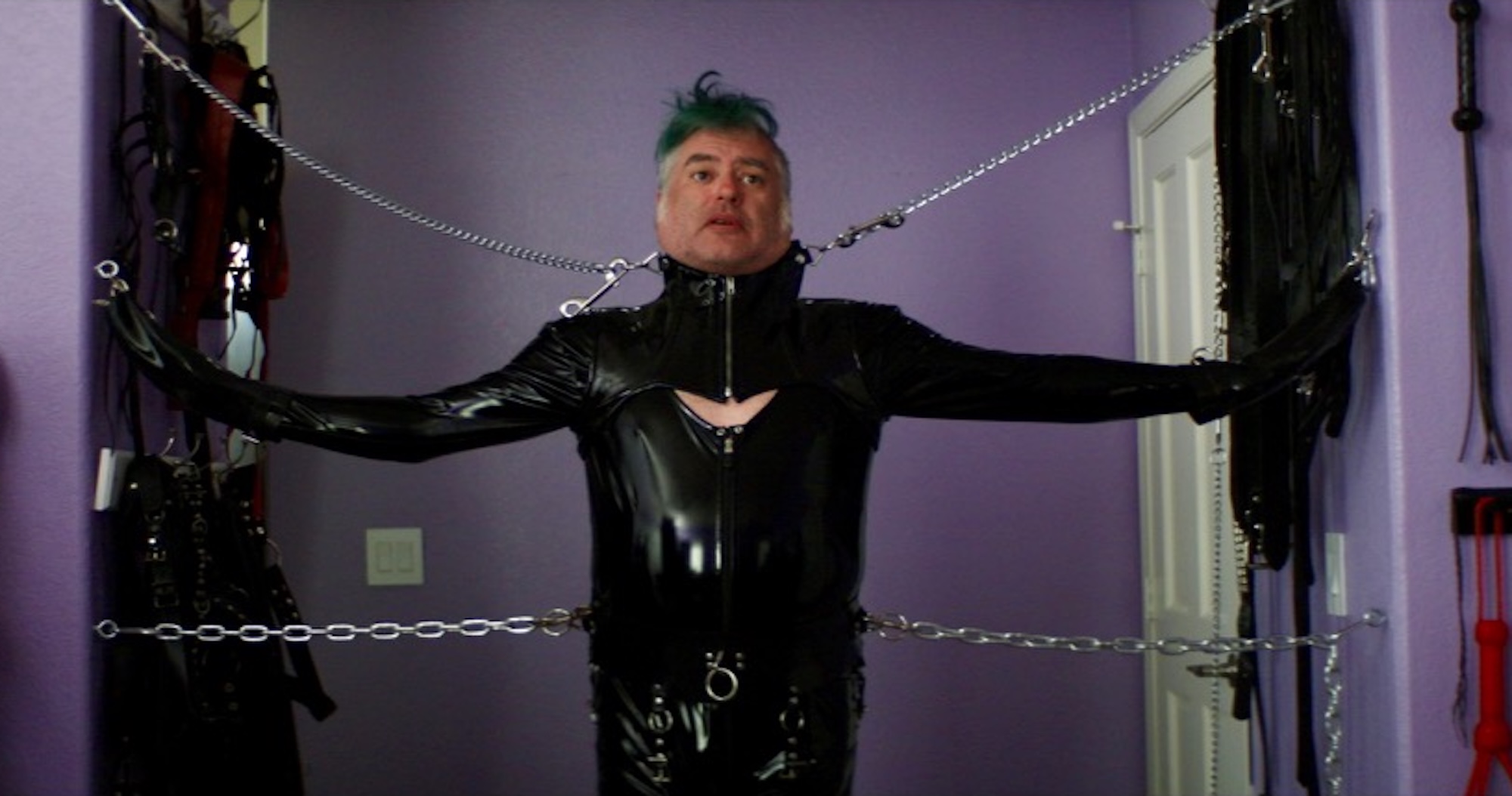 A white man with a green mohawk stands wearing a black rubber suit and dog collar with his arms and neck tethered to the wall via chains.