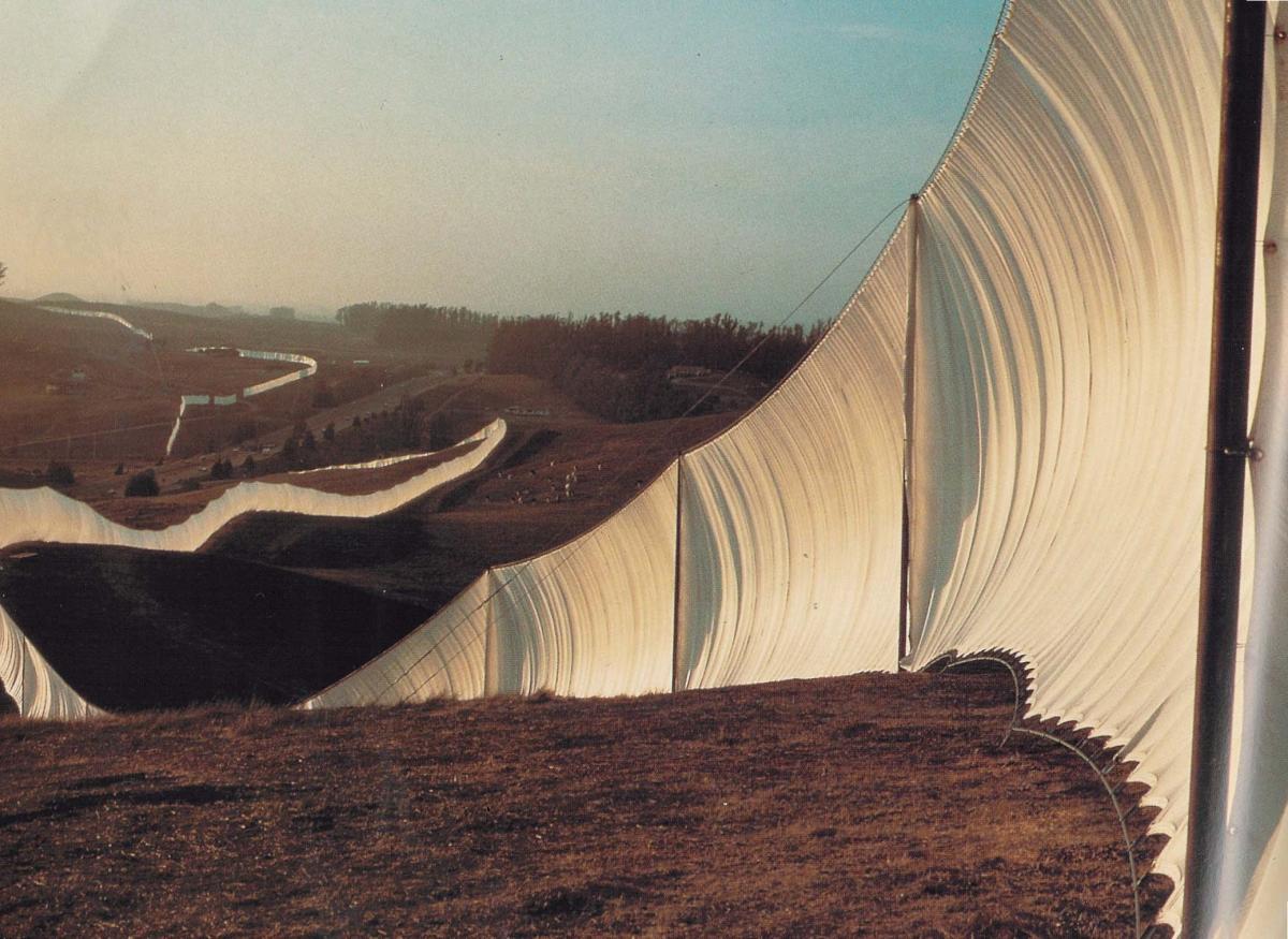 A tall fence made of white fabric snakes across arid farmland hills