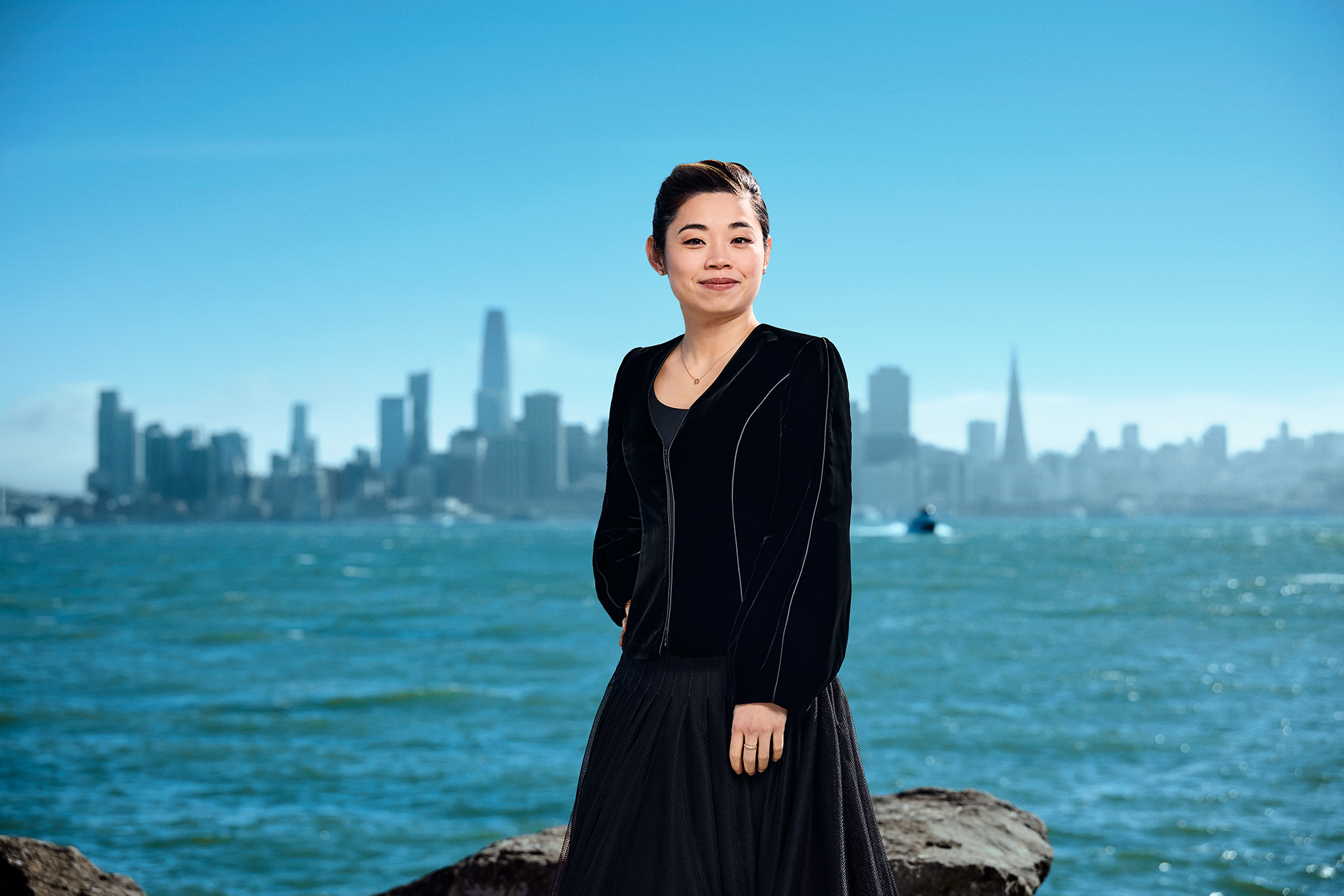 A young smiling Asian woman stands before a body of water with the San Francisco skyline in the background