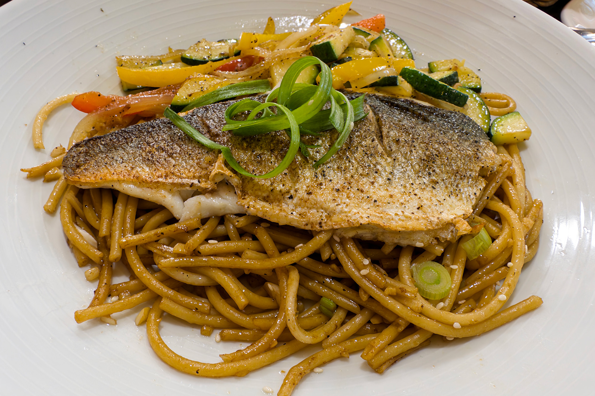 Seared fish fillet on a bed of noodles, with grilled vegetables on the side.