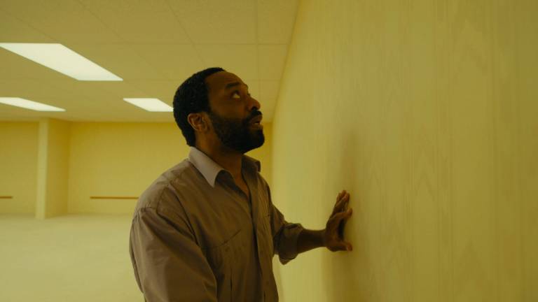 A Black man feels the wall inside an empty yellow-tinted room.