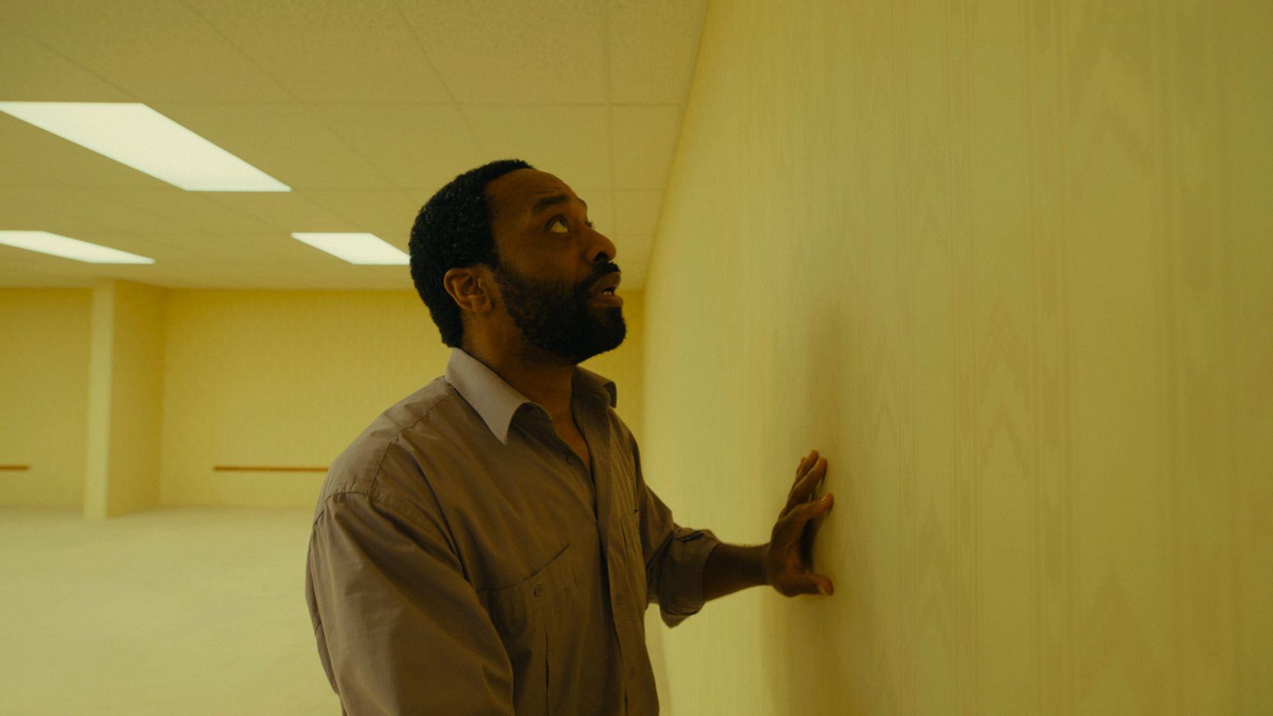 A Black man feels the wall inside an empty yellow-tinted room.