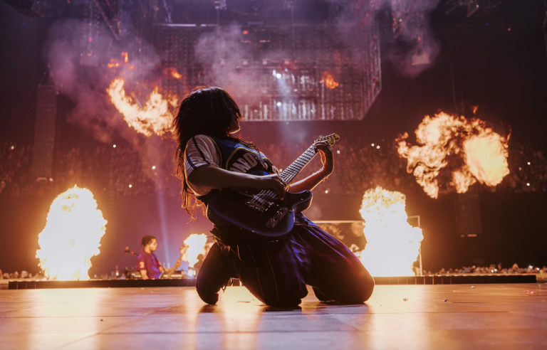 A 20-something woman with long messy hair plays guitar on her knees as flames shoot out of the stage behind her. She is wearing a baggy shirt and pants.