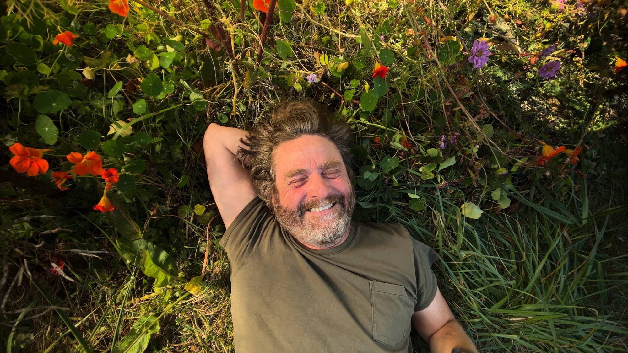 A middle aged bearded white man lies back in the grass, smiling, one arm tucked behind his head.