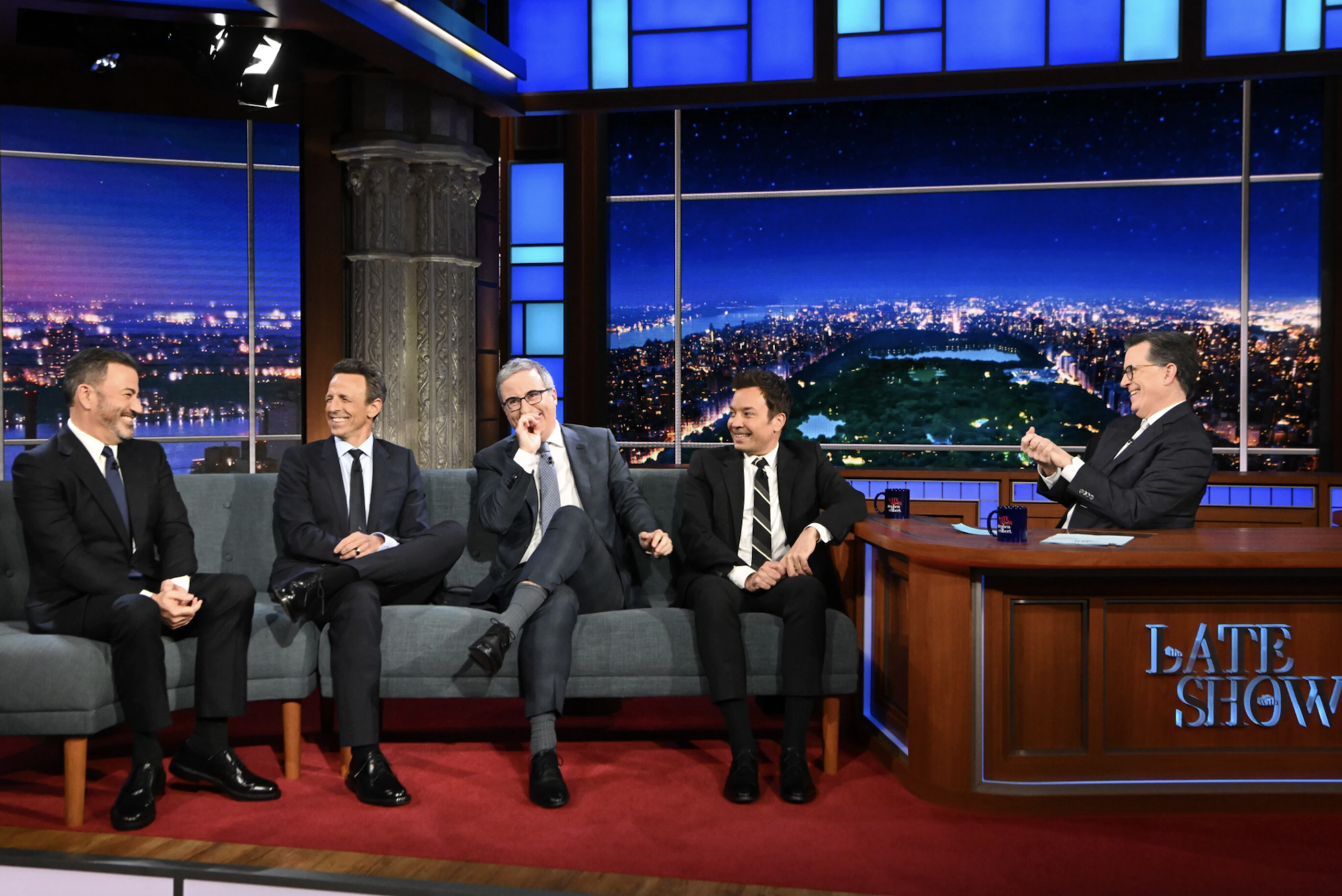 Four middle aged white men in suits laugh together sitting side-by-side on a grey couch in a late night TV studio. To their right, the host - another middle aged white man - claps his hands and laughs from behind a large desk labeled LATE SHOW.