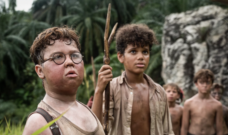 A white boy with round glasses and a mixed race boy holding a spear stand outdoors, close to a rock face and a jungle. They are sweaty and dirty, wearing filthy white shirts.