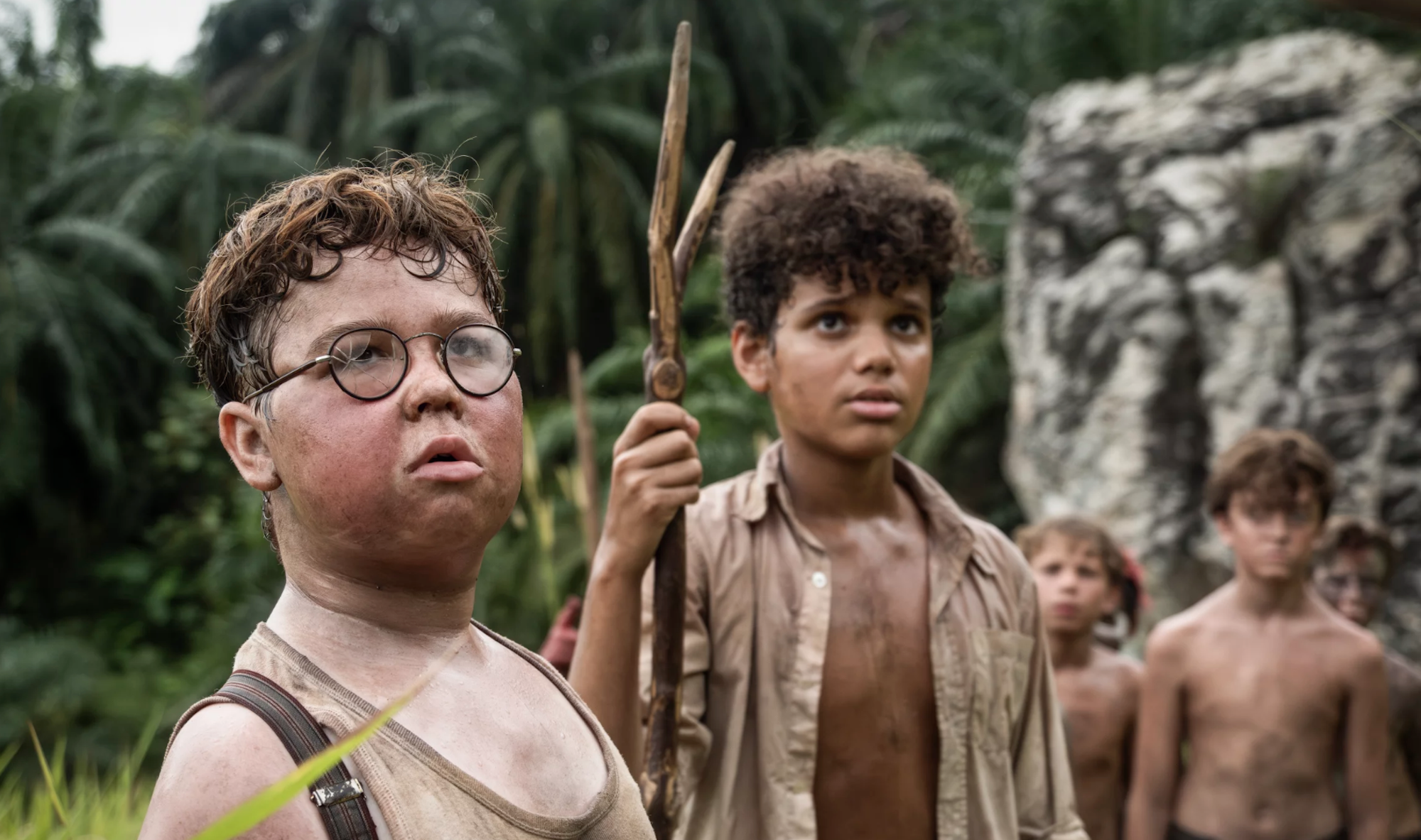 A white boy with round glasses and a mixed race boy holding a spear stand outdoors, close to a rock face and a jungle. They are sweaty and dirty, wearing filthy white shirts.